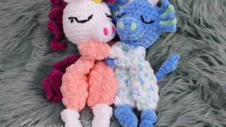 Two plush crochet mini ragdoll loveys, a unicorn and a dragon, snuggle together on a soft green faux fur surface. The unicorn has a white head, pink and peach body, magenta mane, and a yellow horn, while the dragon is blue with green and white accents. Their sleepy embroidered faces add a cozy charm.